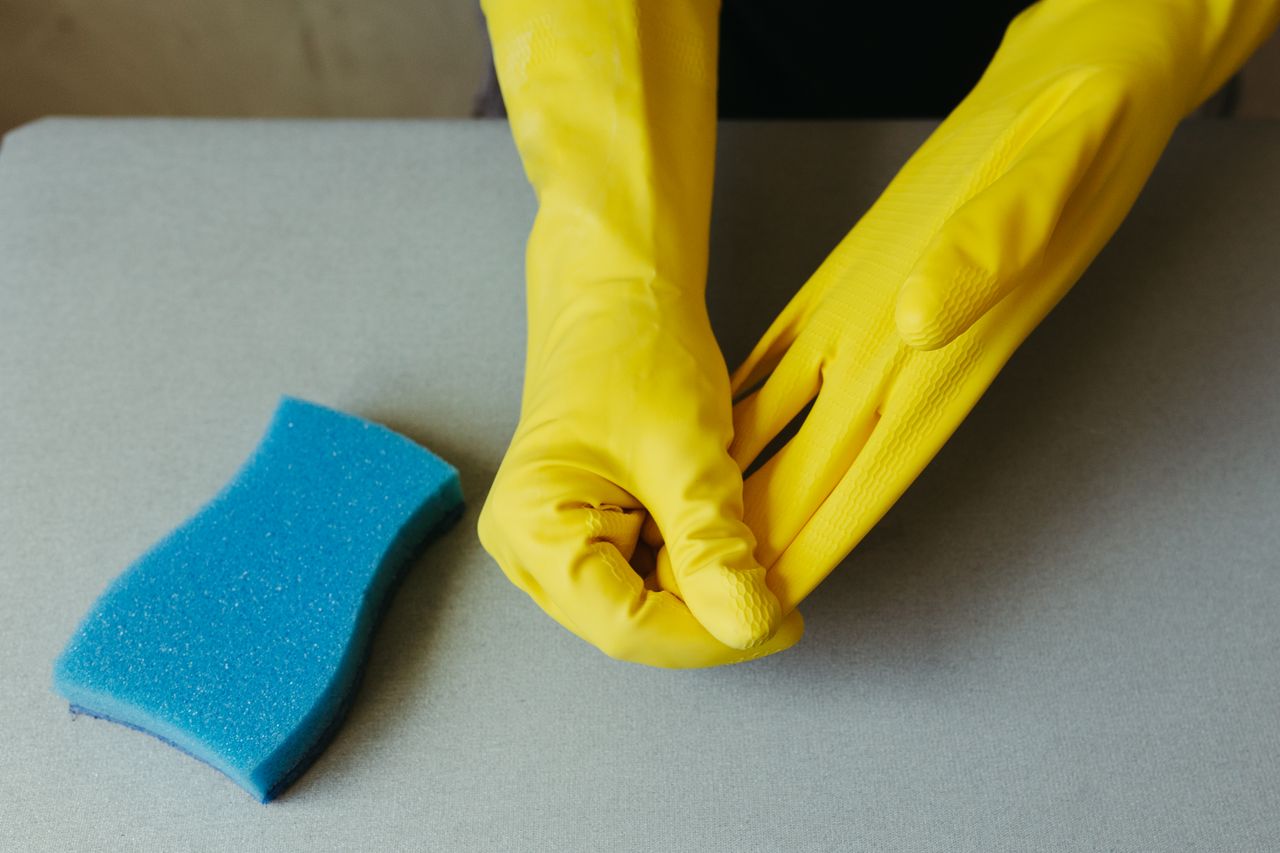Gros plan sur des mains portant des gants jaunes, à côté d’une éponge bleue sur une surface grise.